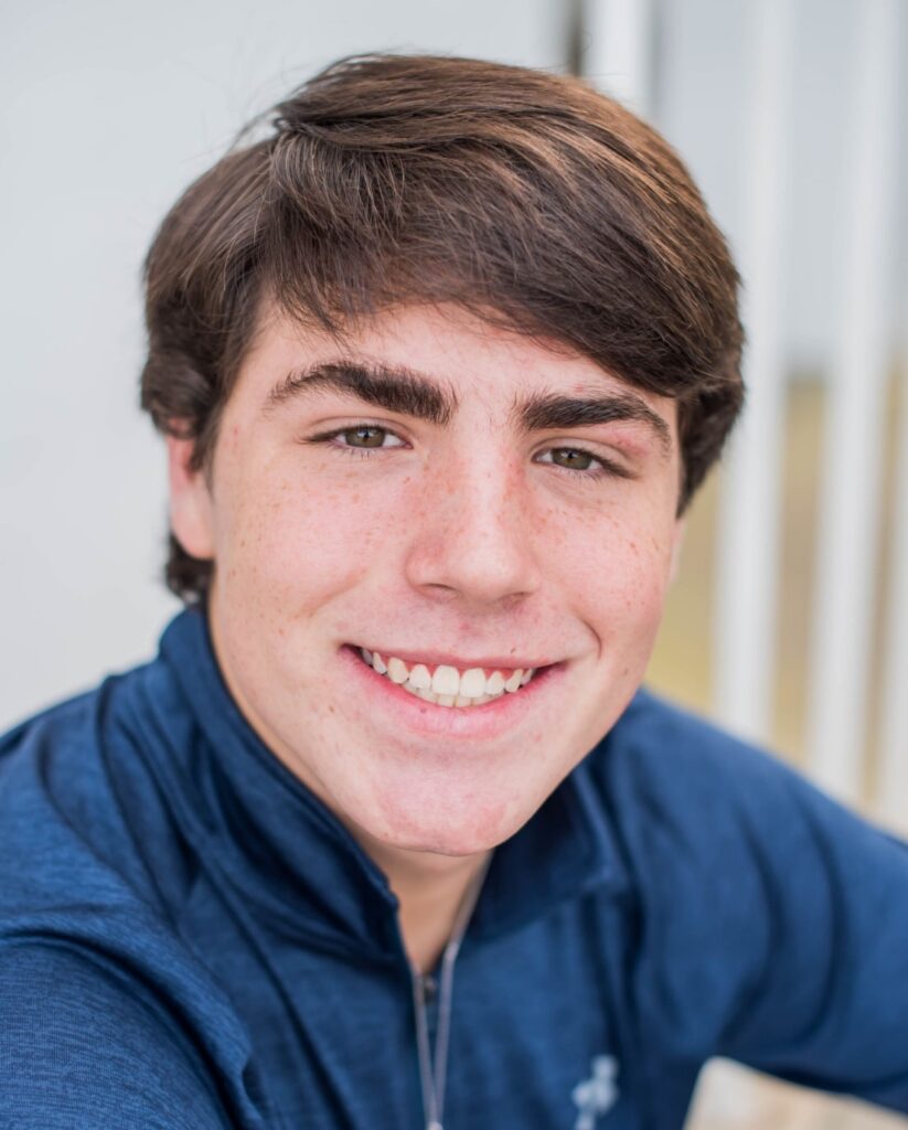 Young teen boy with brown hair and blue zip-up jacket smiling at camera after orthodontic treatment with Morejon + Andrews Orthodontics